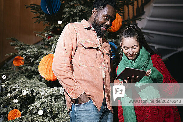 Couple standing near Christmas tree and using tablet PC at home