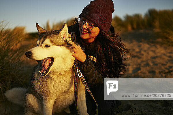 Smiling woman wearing knit hat and petting Husky dog at sunset
