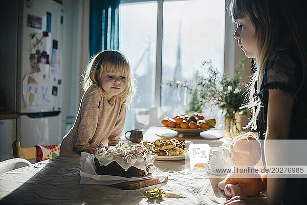 Sisters leaning and looking at cake on table in kitchen
