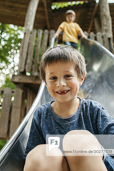 Happy boy sliding down on slide with brother in background at playground