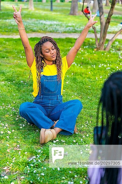 Vertikales Foto einer jungen Afrikanerin in Latzhose  die mit erhobenen Armen sitzt  während ihr Freund ein Foto im Park macht