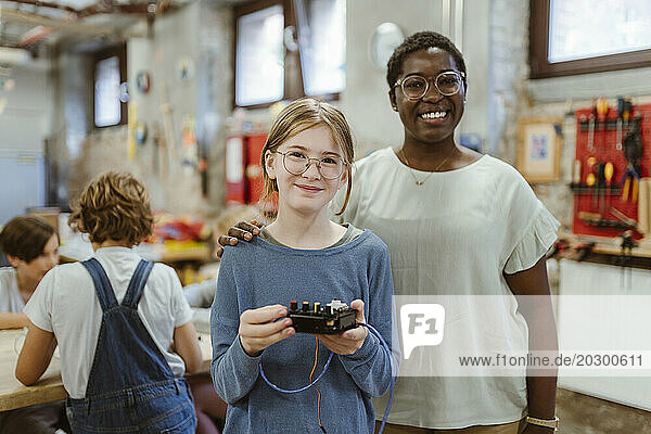 Portrait of smiling teacher with hand on shoulder of female student holding electrical part at technology workshop in sc