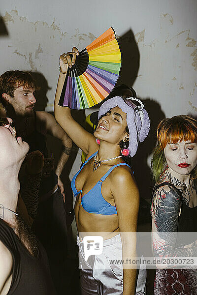 Smiling non-binary person holding multicolored hand fan and dancing with friends at nightclub
