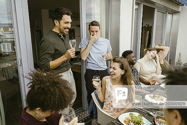 Group of male and female multiracial friends laughing while enjoying drinks at dinner party in balcony