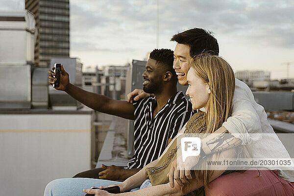 Young man taking selfie with friends through smart phone while sitting on terrace