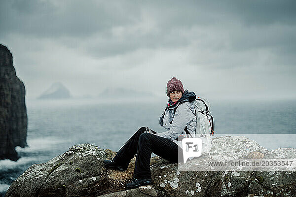 Woman sits on rock while hiking by the ocean in winter Faroe Islands