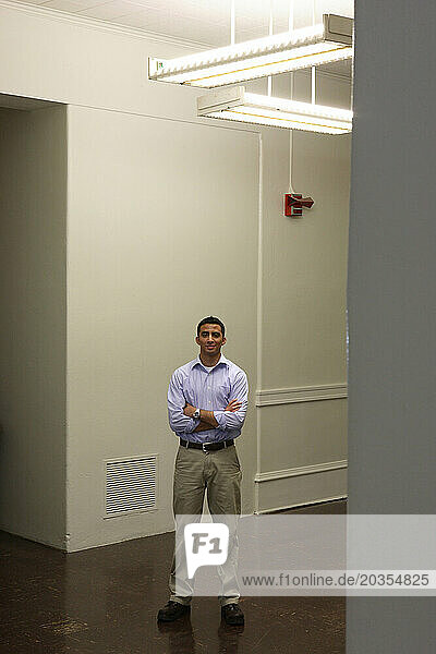 A young hispanic male student stands in a large empty room with his hands crossed.