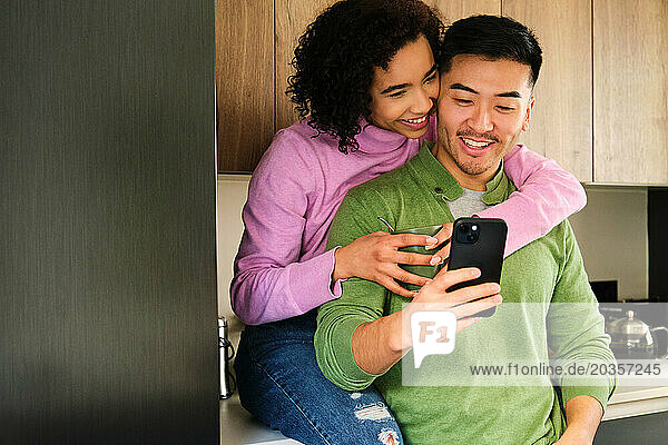 Multiracial couple smile and use the phone together in the kitchen.