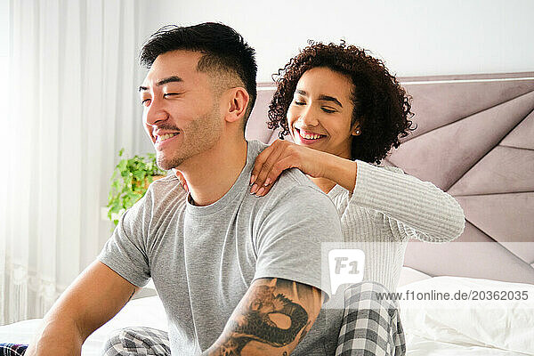 Latin woman massaging her Asian boyfriend shoulders on bed.