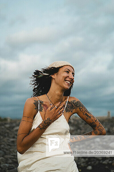 Young happy tattooed woman in a dress on a windy rocky beach.