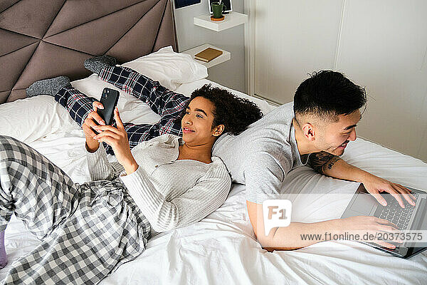 Multiethnic couple smile and use the phone and laptop on bed together.