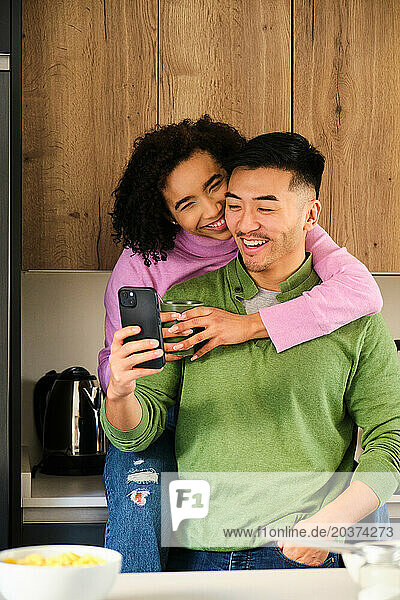 Multiethnic happy couple use the phone and drink coffee in kitchen.