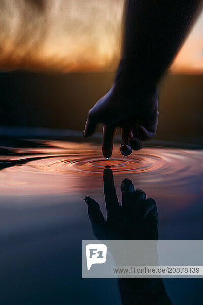 Hand reaching out and touching water with sunset sky