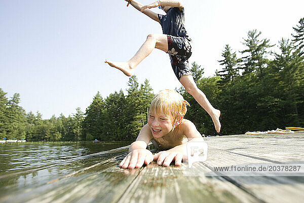 One kid jumps over another into a lake off a wooden raft in Center Harbor  New Hampshire.