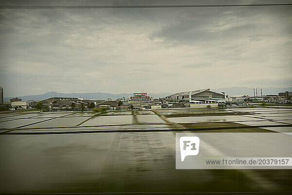A countryside view of farmland an industrial establishment and distant hills from the high-speed Japan bullet train.