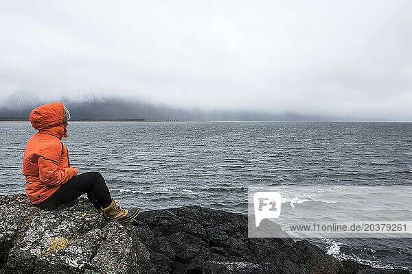 Snowboarder sitting on rock and looking over ocean on stormy day