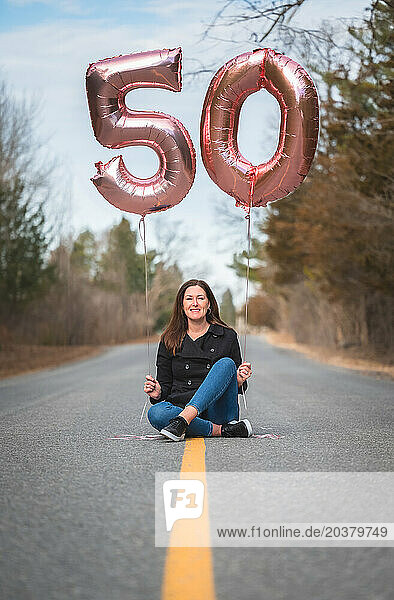 Portrait of woman sitting on road holding age 50 balloons.
