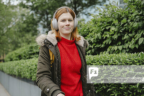 Smiling redhead woman listening to music through wireless headphones near plants