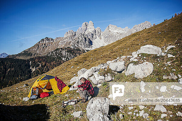 Senior man cooking food near rocks and camping on mountain