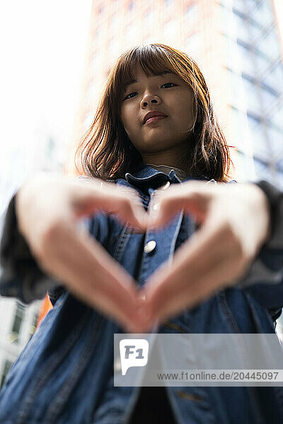 Smiling young woman gesturing heart shape in front of building