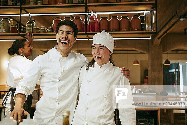 Happy male chef with arm around female chef enjoying while standing in commercial kitchen