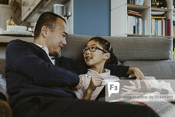 Smiling girl holding digital tablet while sitting with father on sofa at home