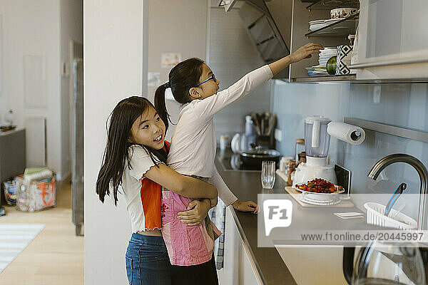 Side view of girl helping sister in reaching at cabinets in kitchen