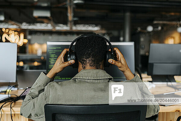 Rear view of male coder wearing wireless headphones sitting at desk in office