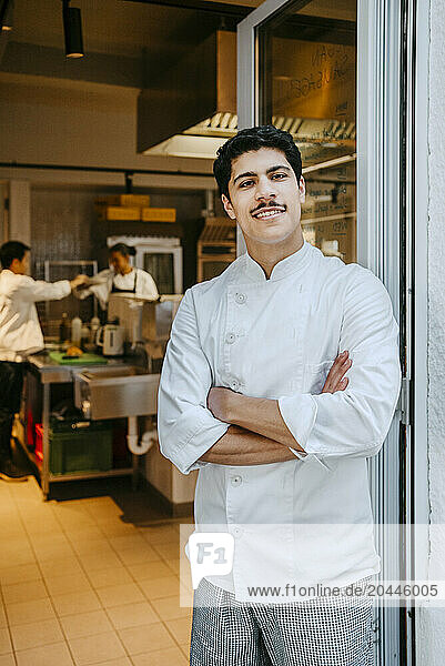 Portrait of happy male chef with arms crossed standing at door in commercial kitchen
