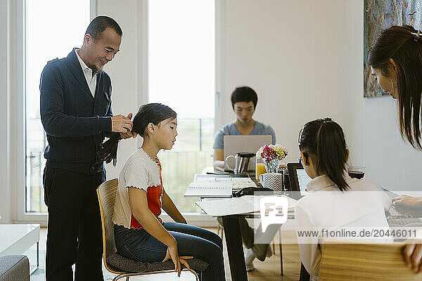 Father tying hair of daughter sitting on chair near dining table at home