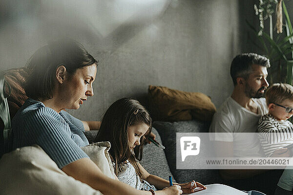 Mother assisting daughter doing homework while sitting on sofa at home