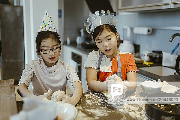 Sisters wearing decorated hats and making dough balls in kitchen at home