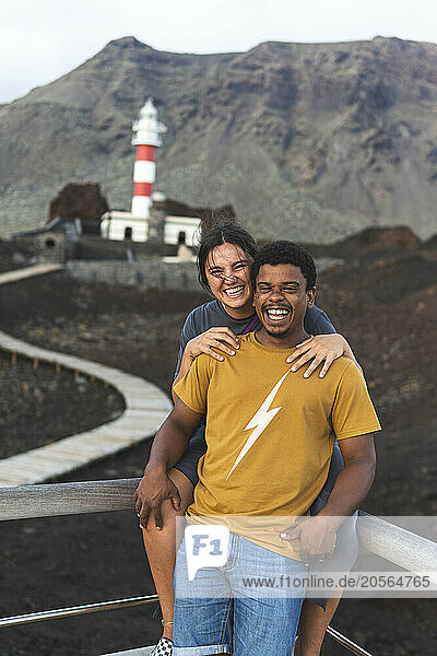 Happy young woman with arm around boyfriend posing in front of Punta Teno Lighthouse