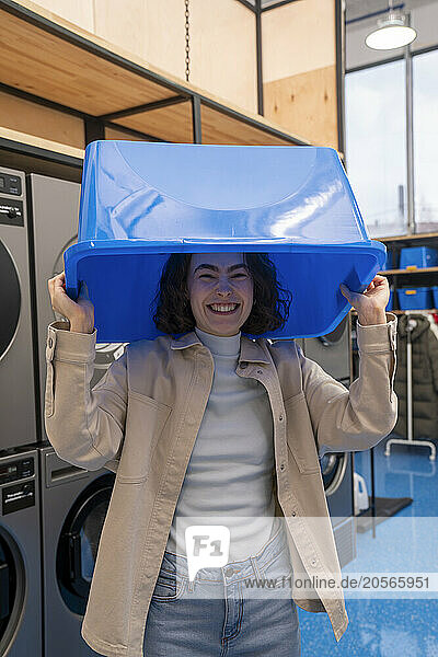 Cheerful young woman with blue basket on top of head standing in laundromat