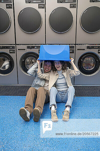 Cheerful young couple with blue basket over head sitting in front of washing machines at laundromat