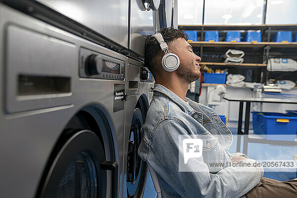Young man listening to music through wireless headphones leaning on washing machine in laundromat