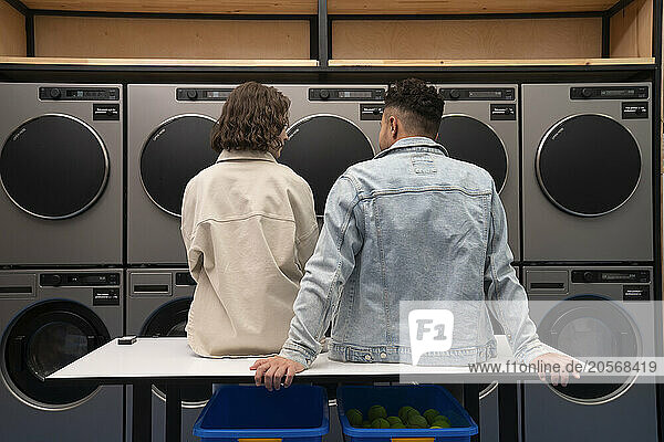 Young boyfriend and girlfriend sitting on table in front of washing machines at laundromat