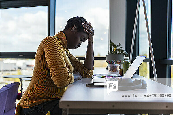 Stressed businesswoman with hand on head and sitting near desk at office