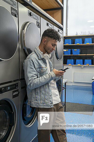 Young man leaning on washing machine using smart phone in laundromat