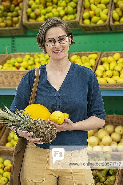 Smiling mature woman in eyeglasses carrying fruits and standing in supermarket