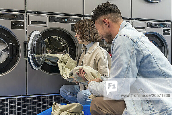 Smiling young couple removing sheet from washing machine at laundromat