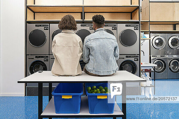 Young couple sitting on table in front of washing machines at laundromat