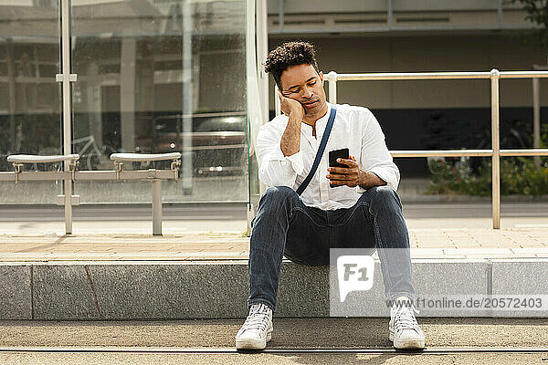 Man leaning on elbow and using smart phone sitting at bus stop