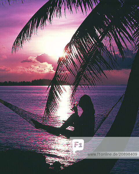 Guam. Tropical sunset silhouette of young woman sitting in hammock with glass of wine.