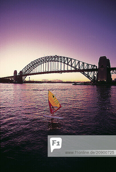 Australia. Sydney. Harbour Bridge with young woman sailboarder at sunset.