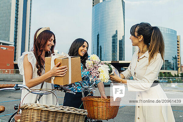 Happy woman giving gifts and bouquet of flowers to friend in front of buildings