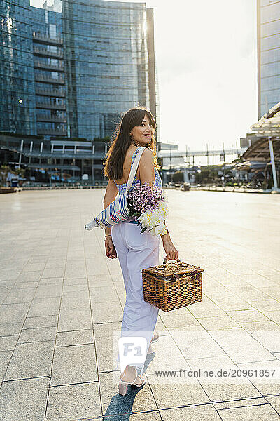Smiling woman walking with bouquet of flowers and basket on footpath