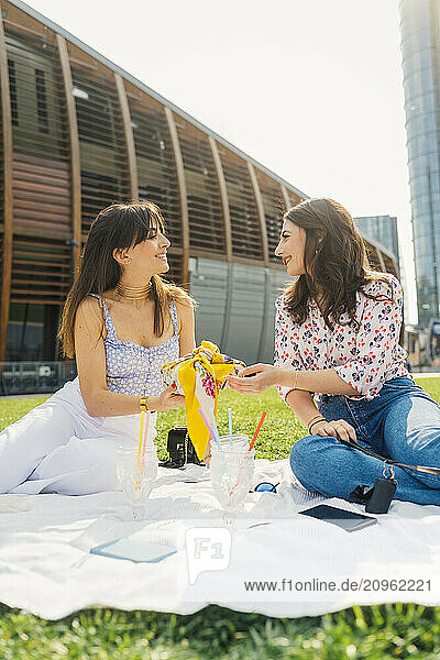 Smiling woman giving gift to friend sitting on picnic blanket