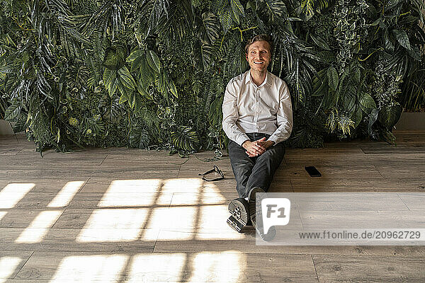 Happy businessman sitting on floor in front of plants