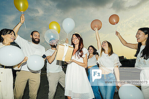 Happy friends celebrating woman's birthday with balloons and gift at sunset